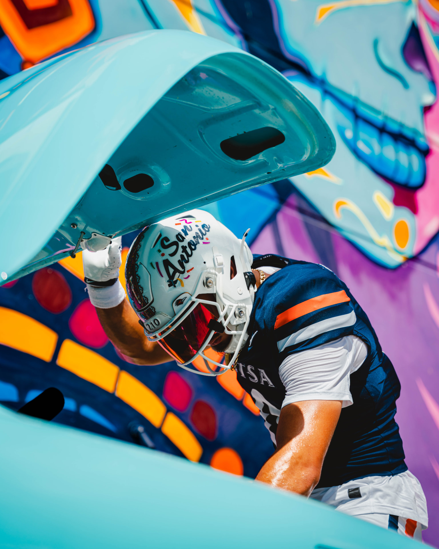 UTSA player wearing the San Antonio helmet beside a turquoise car and colorful mural